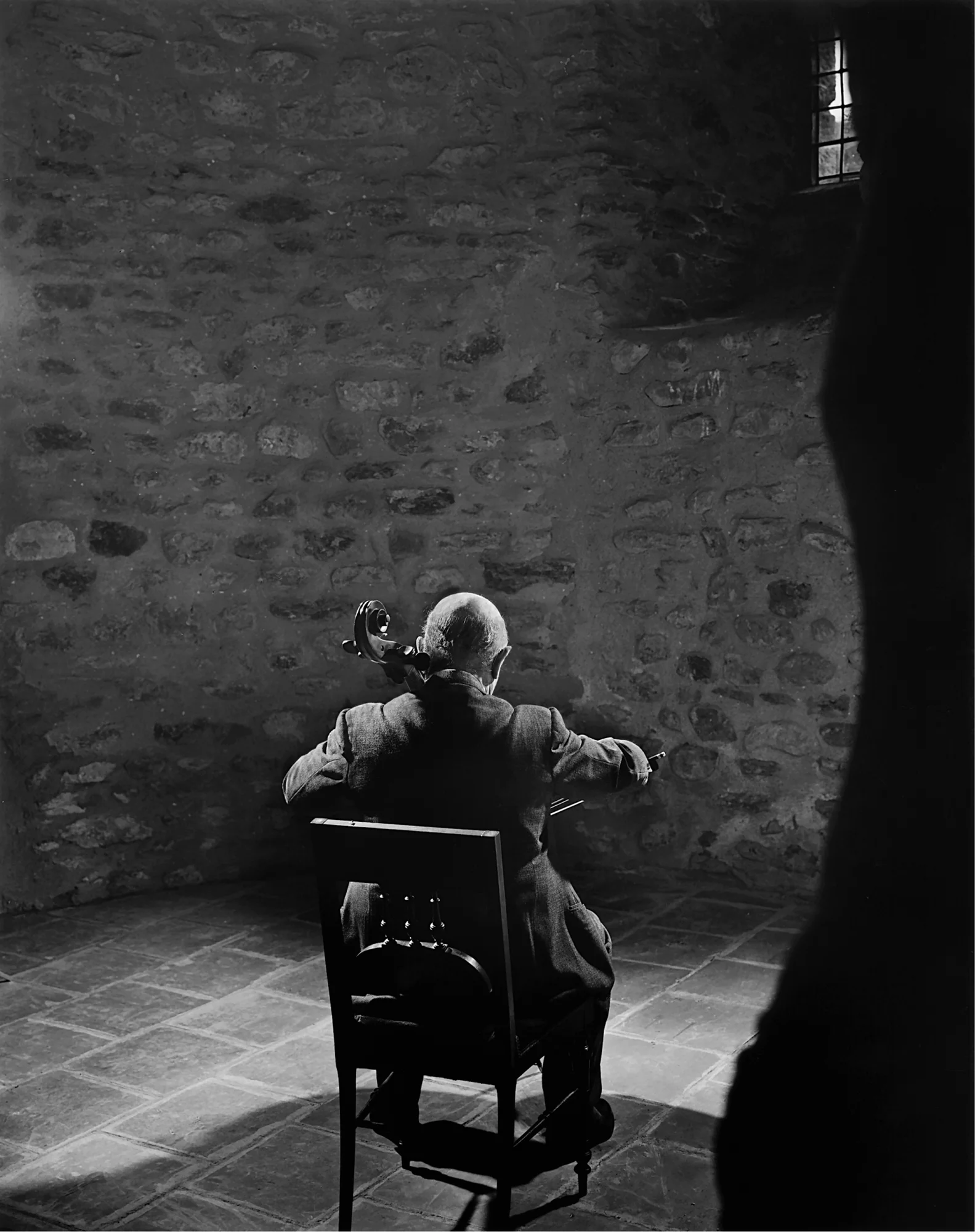 Pablo Casals seated on a chair facing away from the viewer, playing a cello.