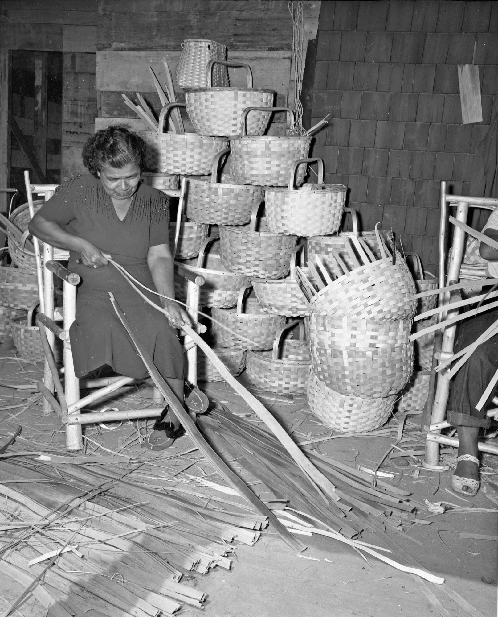 Black-and-white photo of a woman sitting and weaving wooden strips into baskets, with many finished baskets stacked behind her.